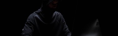 panoramic shot of hooded hacker in mask on black  clipart