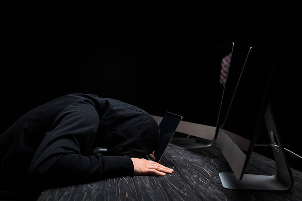 upset hacker lying on laptop near computer monitors with blank screen isolated on black 