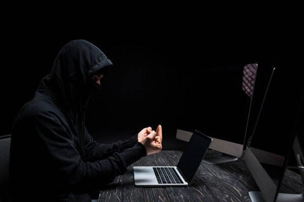 displeased hacker showing middle finger near laptop and computer monitors with blank screen isolated on black 