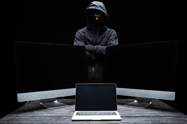 hacker in mask standing with crossed arms near computer monitors and laptop with blank screen  isolated on black 