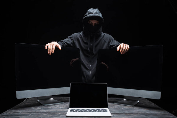hacker in mask touching computer monitors near laptop with blank screen isolated on black 