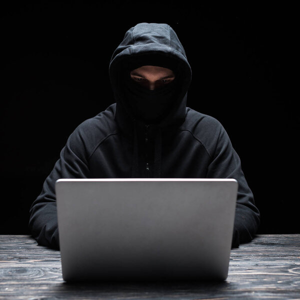 молодой хакер в маске и капюшоне с помощью ноутбука изолированы на черный
 