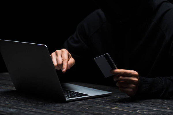 cropped view of hacker using laptop while holding credit card isolated on black 