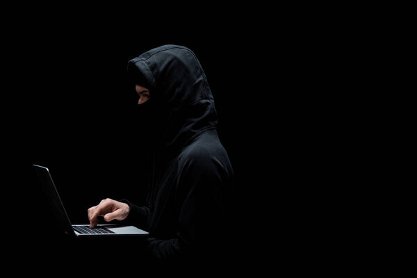 anonymous hacker in hood and mask using laptop isolated on black 