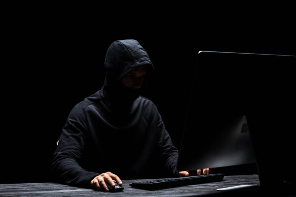 hooded anonymous hacker in mask using computer isolated on black 