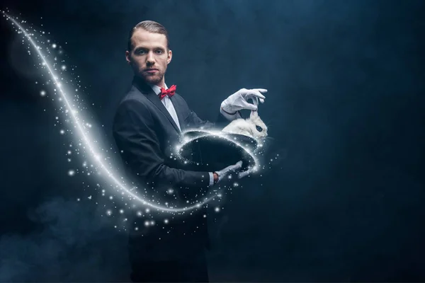 スーツ姿のプロの魔術師が白いウサギを帽子に煙と輝くイラストの暗い部屋で — ストック写真