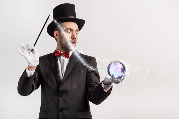 shocked magician holding wand and magic ball isolated on grey with glowing illustration