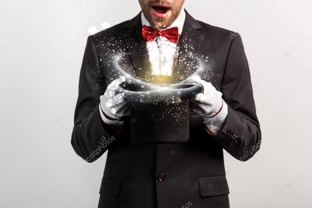 cropped view of shocked magician holding hat isolated on grey with glowing illustration