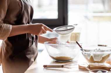 cropped view of young woman sieving flour in glass bowl near whisk  clipart