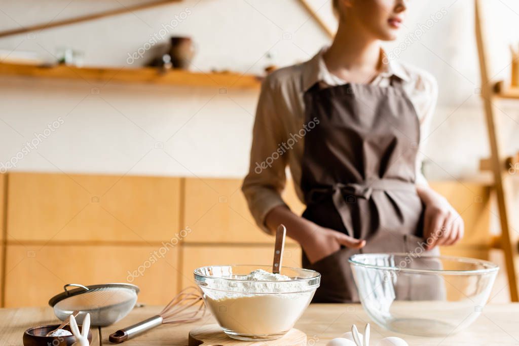 cropped view of woman in apron standing with hand in pocket near bowls and ingredients 