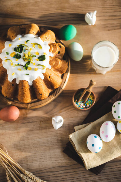 вид сверху на пасхальный торт возле стакана молока, статуэтки с кроликами и покрашенные яйца
 
