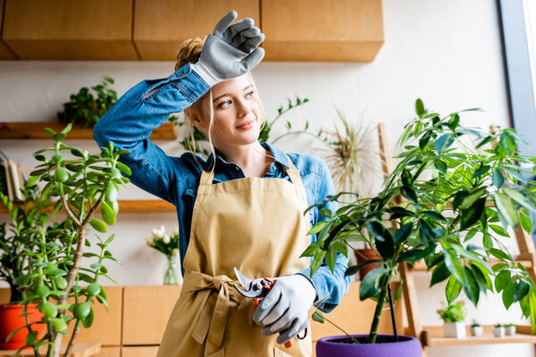 tired young woman in gloves smiling while holding gardening scissors near plants 
