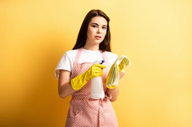 Önlüklü, lastik eldivenli, sarı arka planda deterjan sıkan düşünceli bir ev hanımı.