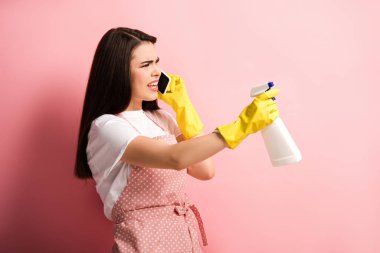 Önlüklü ve lastik eldivenli hoşnutsuz ev hanımı elinde pembe arka planda sprey şişeyle akıllı telefondan konuşuyor.