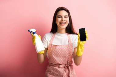 Önlüklü ve lastik eldivenli mutlu ev hanımı pembe arka planda sprey şişeyi tutarken boş ekranlı akıllı telefonu gösteriyor.