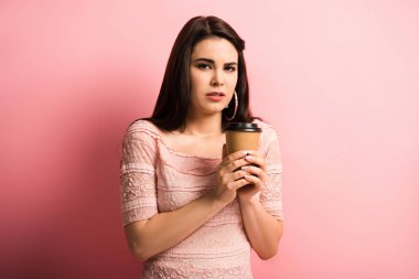 Pembe arka plandaki kameraya bakarken elinde kahve tutan rahatsız bir kız.