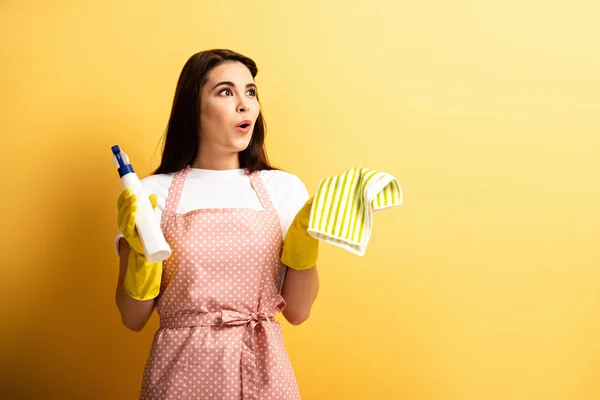 surprised housewife in apron and rubber gloves holding spray bottle and rag on yellow background