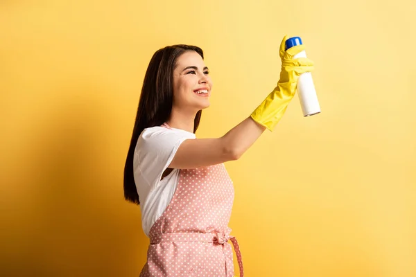 cheerful housewife in apron and rubber gloves spraying air freshener on yellow background