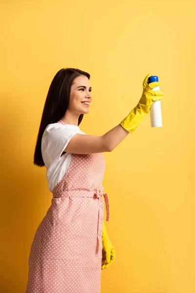 happy housewife in apron and rubber gloves spraying air freshener on yellow background