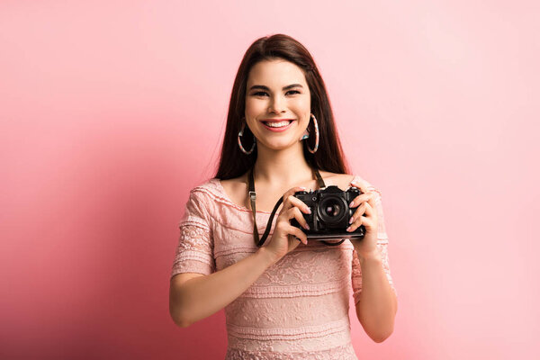 happy photographer smiling while holding digital camera on pink background