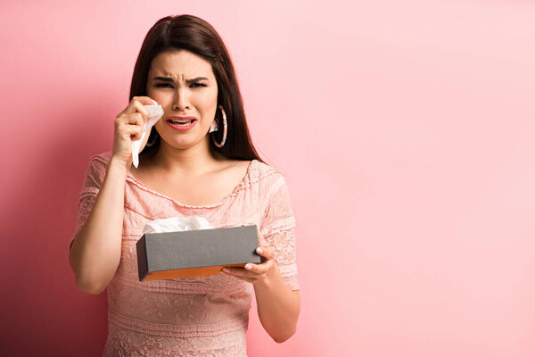 offended girl crying and wiping tears with paper napkin while looking at camera on pink background