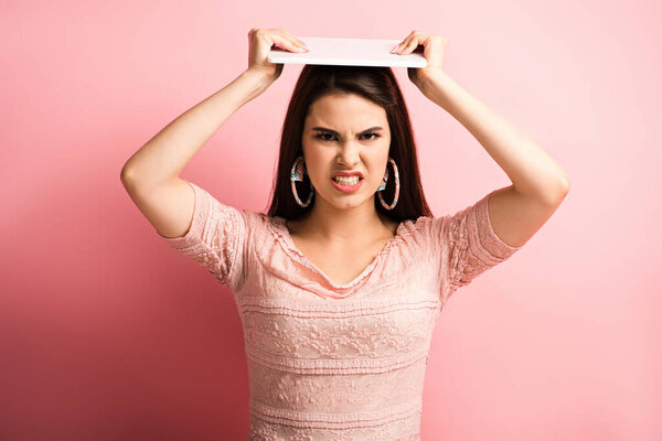 сердитая девушка гримасничает и держит цифровой планшет над головой, глядя на камеру на розовом фоне
