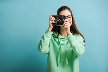 Yeşil kapüşonlu gülümseyen fotoğrafçı mavi arka plandaki dijital kamerada fotoğraf çekiyor.