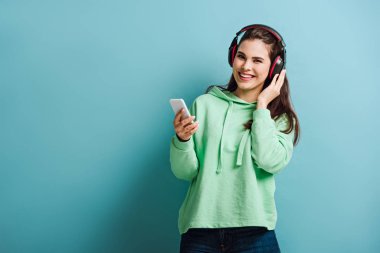 Kablosuz kulaklıklı neşeli kız mavi arka planda akıllı telefonu tutarken kameraya bakıyor.