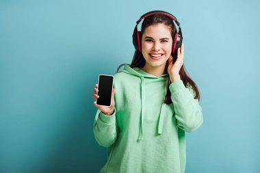 Kablosuz kulaklıklı neşeli kız kameraya bakarken mavi arka planda boş ekranlı akıllı telefonu gösteriyor.