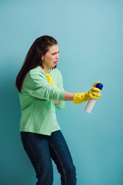 Hoşnut olmayan ev hanımı, mavi arka plana oda spreyi sıkarken yüzünü buruşturuyor.