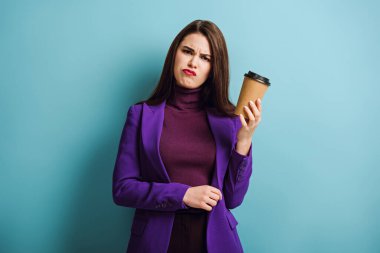 sceptical girl looking at camera while holding coffee to go on blue background clipart