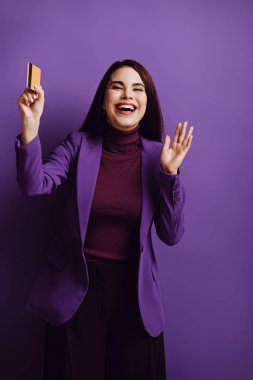 Heyecanlı genç bir kadın mor arka planda kredi kartını tutarken el sallıyor ve gülüyor.