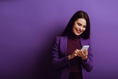cheerful young woman smiling while chatting on smartphone on purple background clipart