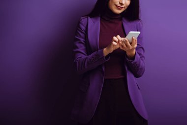 cropped view of stylish woman typing on smartphone on purple background clipart