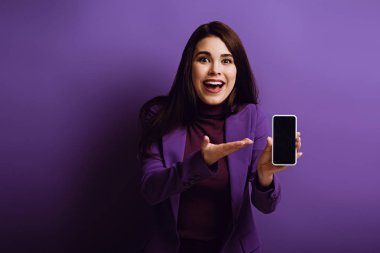 Mor arka planda boş ekran ile akıllı telefonu işaret eden sürpriz kız