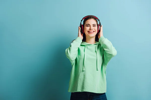 Happy Girl Touching Wireless Headphones While Looking Camera Blue Background — Stock Photo, Image