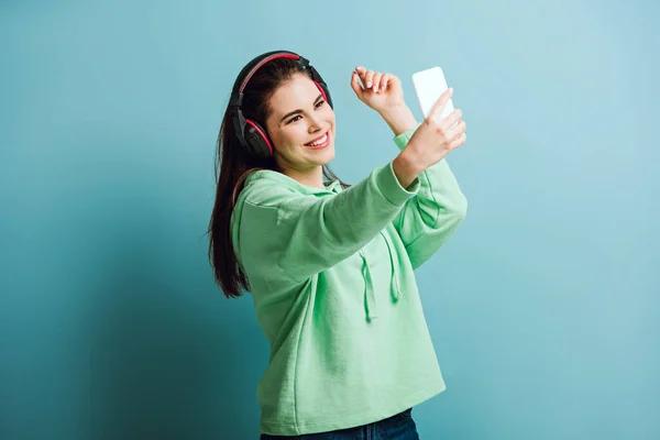 happy girl in wireless headphones taking selfie on smartphone on blue background