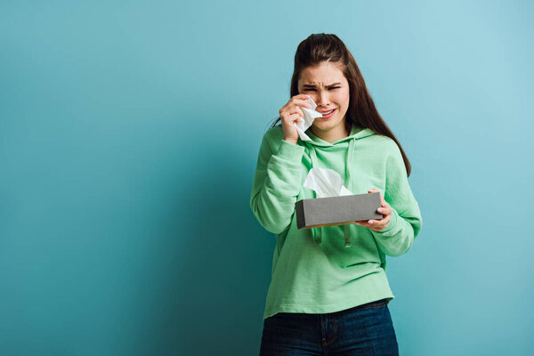 upset, crying girl looking at camera while wiping tears with paper napkin on blue background