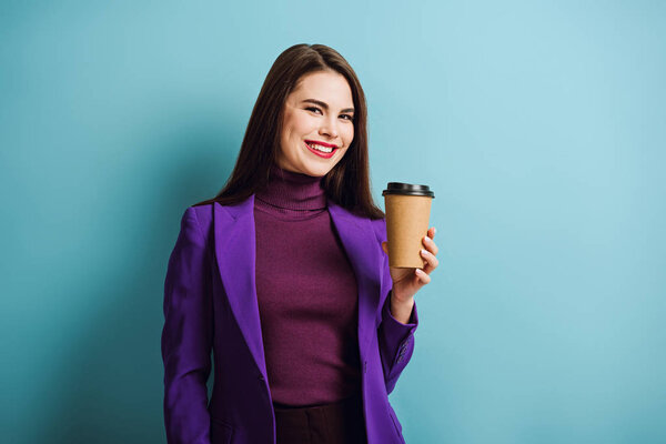 cheerful girl looking at camera while holding coffee to go on blue background