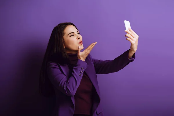 happy young woman blowing air kiss while taking selfie on purple background