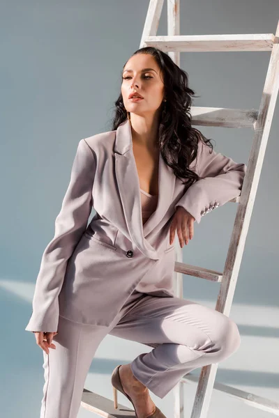 stylish woman in suit near ladder looking away on grey background