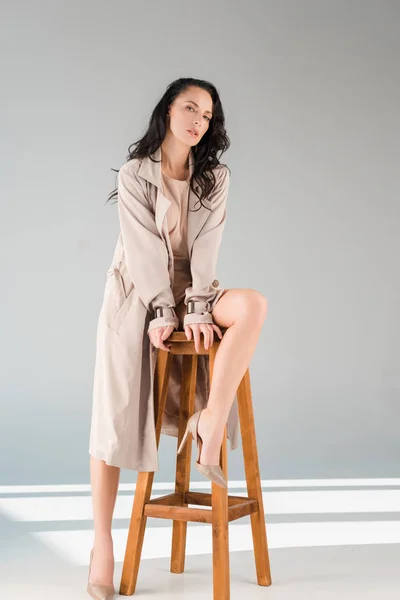 attractive woman in coat sitting on stool on grey background