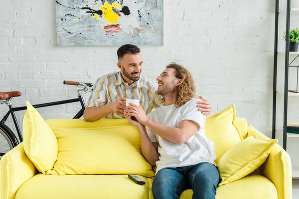 cheerful homosexual men smiling while holding cup of tea at home 