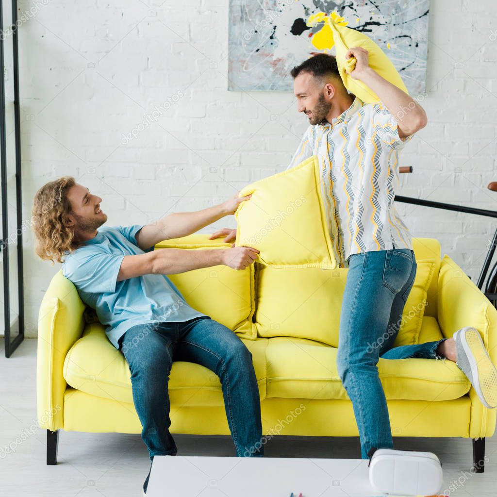 Side view of cheerful homosexual men pillow fighting in living room