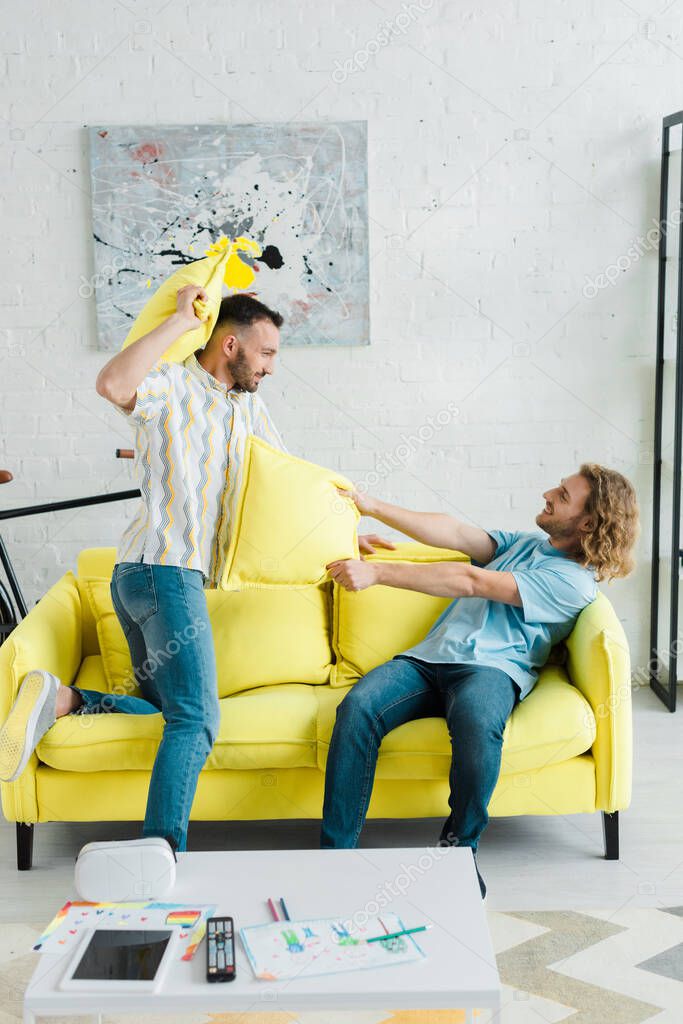 Profile of happy homosexual men pillow fighting in living room