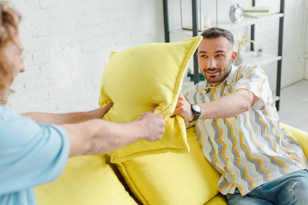 Selective focus of cheerful homosexual men pillow fighting in living room
