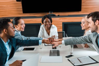 happy, young multicultural businesspeople holding joined hands while sitting at desk in conference hall clipart