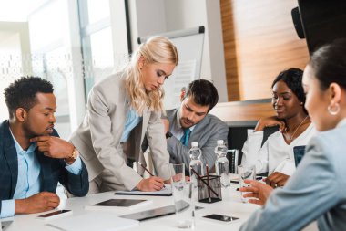 selective focus of attractive businesswoman writing in notebook near multicultural colleagues sitting at desk in conference hall clipart