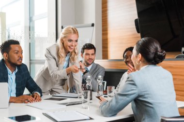 young smiling businesswoman pointing with pencil near multicultural colleagues sitting at desk in conference hall clipart