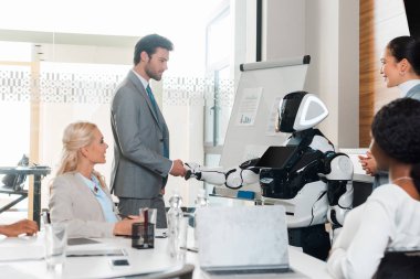 Yakışıklı iş adamı, konferans salonunda oturan çok kültürlü meslektaşlarının yanında robotla el sıkışıyor.